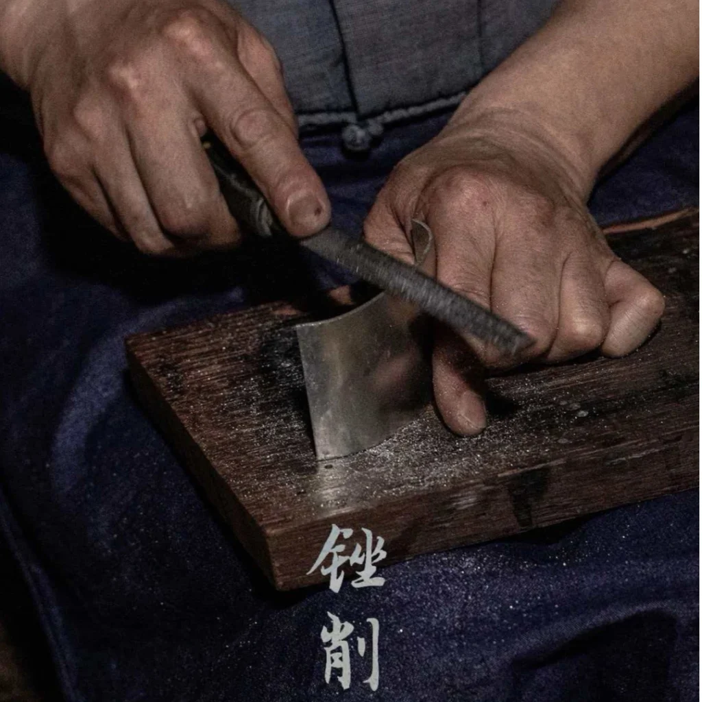 Artisan filing and shaping tin material at workbench during Laiwu tin carving process, metal shavings and hand tools visible, by Heritage & Craft.